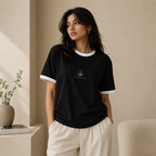 Young Indian woman wearing the Zenrola black unisex ringer T-shirt with white ribbed collar and sleeve cuffs, paired with cream trousers in a warm minimalist studio.