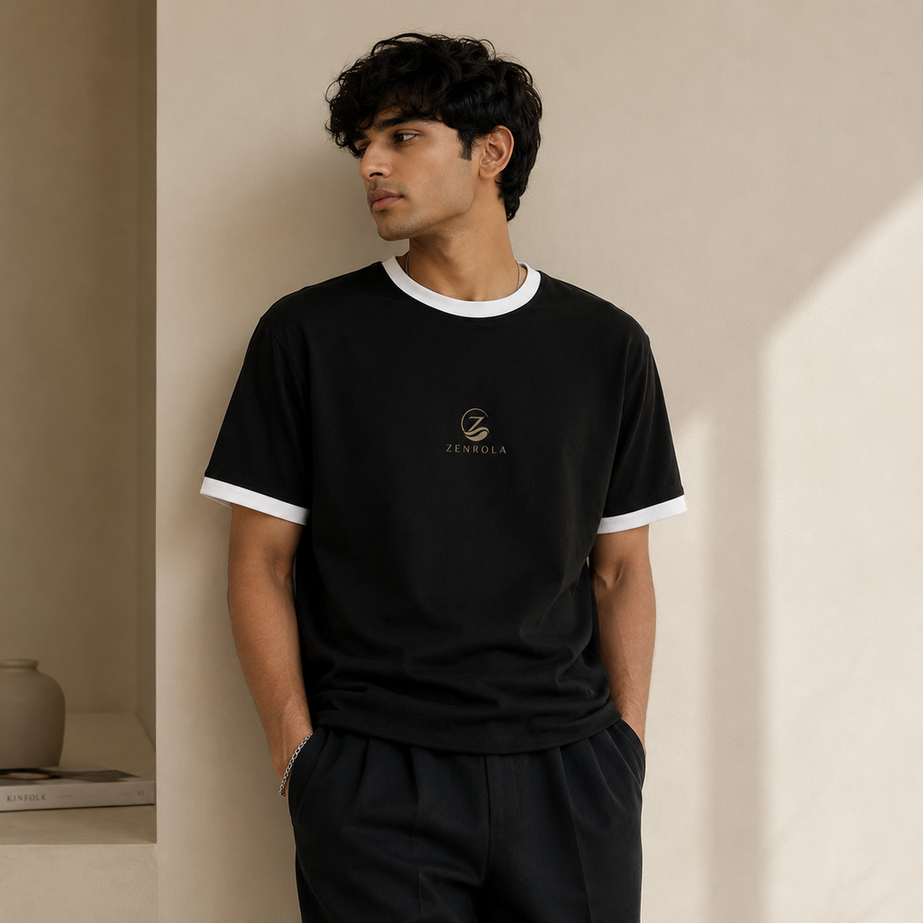 Young Indian man wearing the Zenrola black unisex ringer T-shirt with white ribbed collar and sleeve cuffs, styled with black trousers in a modern minimalist studio.