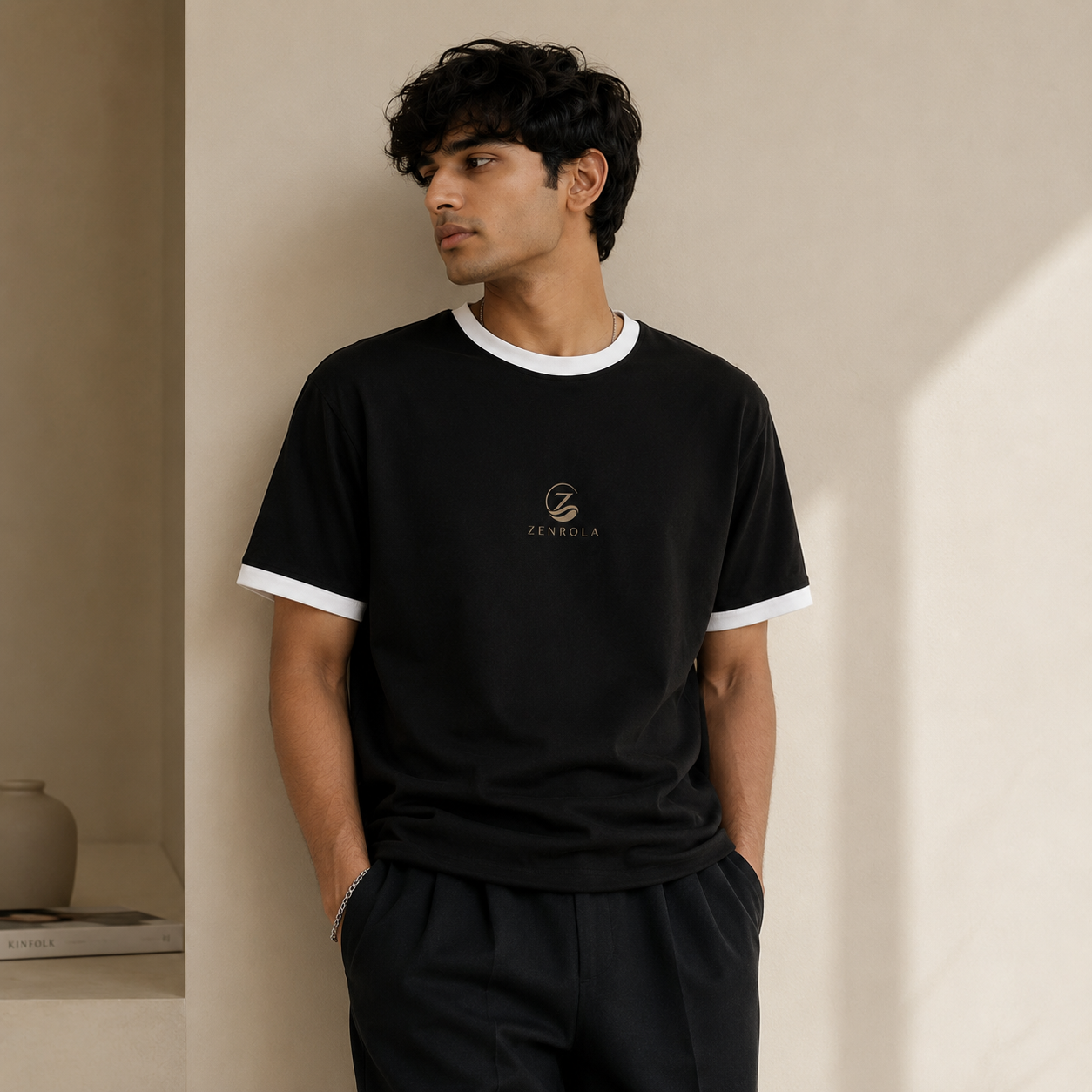 Young Indian man wearing the Zenrola black unisex ringer T-shirt with white ribbed collar and sleeve cuffs, styled with black trousers in a modern minimalist studio.