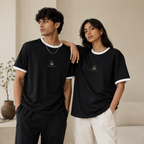 Young Indian man and woman wearing the Zenrola black unisex ringer T-shirt with white ribbed collar and sleeve cuffs in a minimalist beige studio, showcasing a relaxed everyday fit.